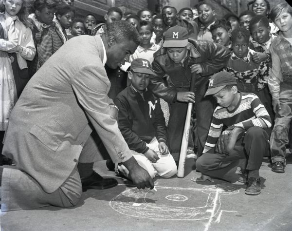 Jackie Robinson crouching on the ground and pointing to a diagram of a baseball diamond sketched on the pavement as a large group of primarily African-American boys, some in Milwaukee Braves caps, are looking on.