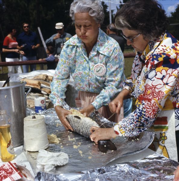 Two elderly women are stuffing and tying a musky with string before cooking it. The women are wearing buttons with an image of a musky that reads: "I am a Boulder Junction Rooster, 'Musky Capital of the World.'" A group of people are watching from the sidelines.