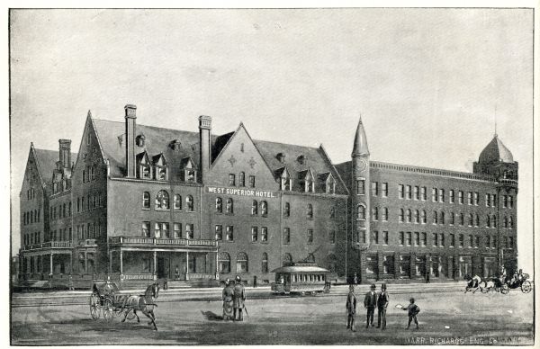 Exterior view of the West Superior Hotel. There are pedestrians, a street car, and people riding in horse-drawn carriages on the street in front of the hotel.
