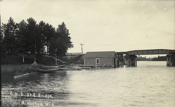 View from water of the Chicago Milwaukee and St. Paul railroad bridge. Probably over a stretch of Kawaguasaga Lake. There is a stairway leading to the water at left. A boathouse and several boats and canoes are along the shoreline. Caption reads: "C.M. & St. P. Bridge, Minocqua, Wis."
