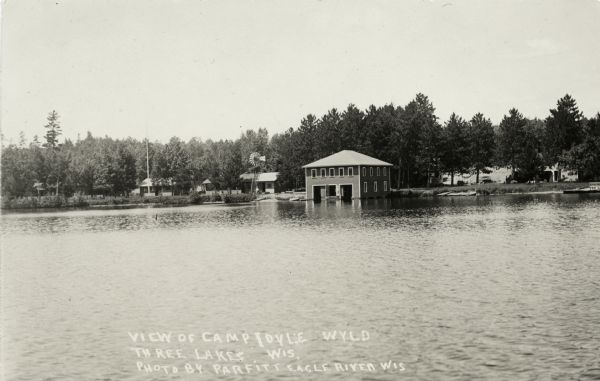 View from lake of Camp Idyle Wyld. A two-story wet boathouse, a windmill, and several cabins are visible along the shoreline.
