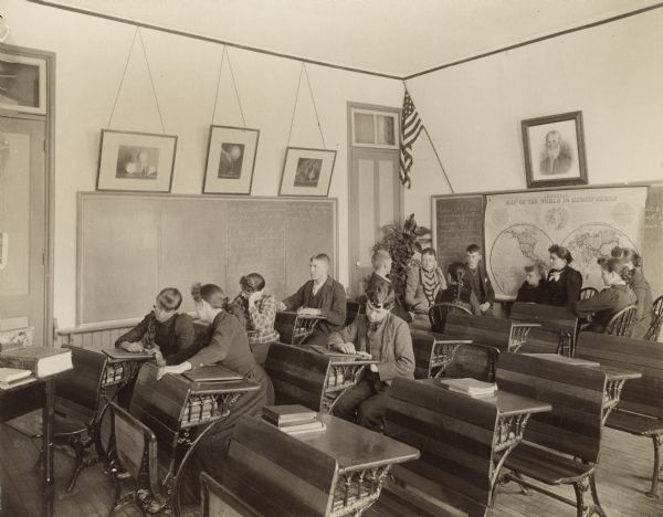 Male and female students sitting in desks while writing on what appear to be slates. A group sitting in a circle at the back of the room are possibly practicing conversation skills.