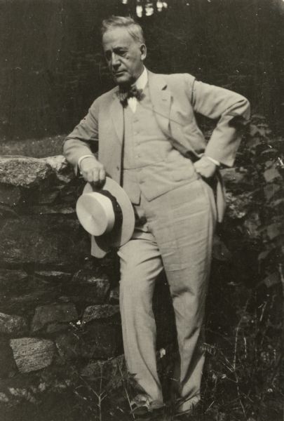 Informal full-length outdoor portrait of Magnus Swenson, wearing a suit and bow tie with a hat in his hand, leaning against a stone wall.
