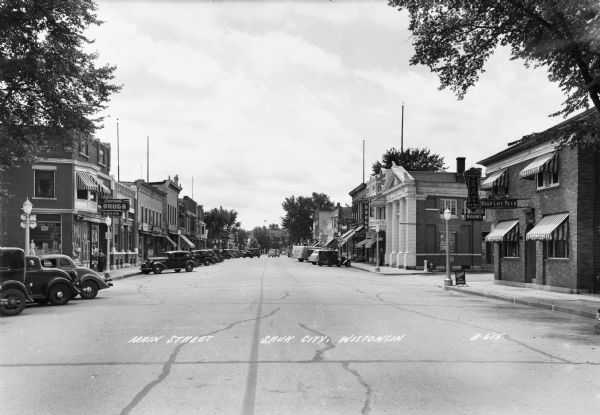 View down the center of Main Street, with many diagonally parked automobiles. Prominent on the right hand side of the street at a corner is the Classic Revival State Bank built in 1913.