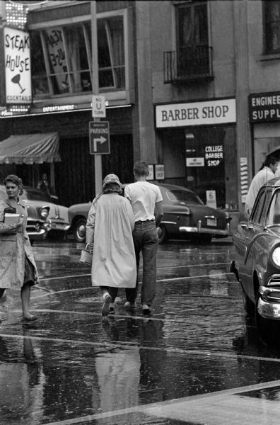 People cross a State Street intersection in the rain. The College Barber Shop and a Steak House is on the other side of the street.