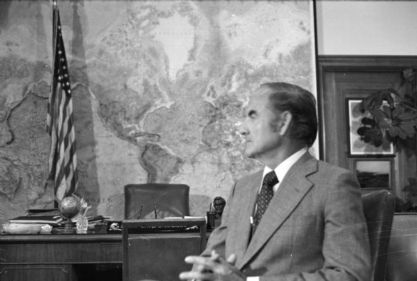 George McGovern photographed during an interview by Robert Novak. Behind McGovern is a desk, American flag, and a large map of the world.