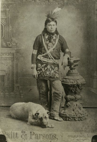 Full-length studio portrait in front of a painted backdrop of a Winnebago (Ho-Chunk) man wearing a headdress. A dog is laying on the floor near his feet.