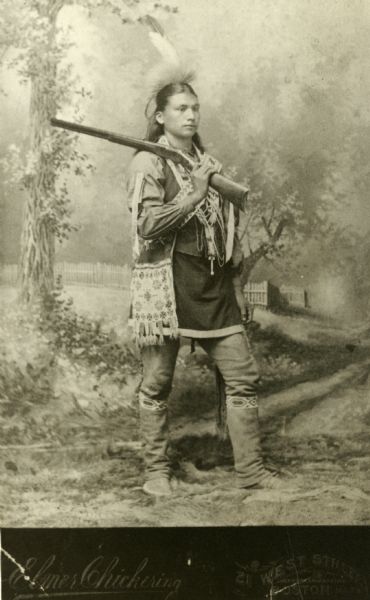 Full-length studio portrait in front of a painted backdrop of a Winnebago (Ho-Chunk) man posed with gun over his shoulder. He carries a beaded bag and wears a headdress.