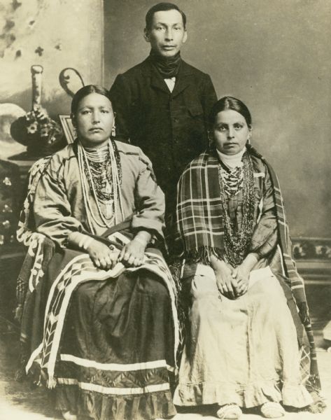 Studio portrait in front of a painted backdrop of a Winnebago (Ho-Chunk) family, including two women sitting and one man standing.