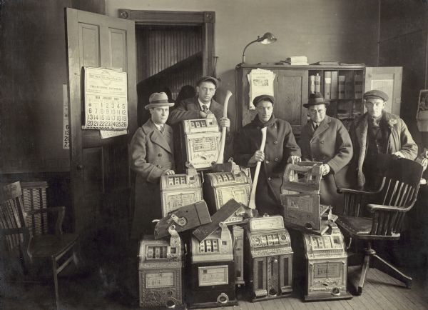 Gerald J. Boileau, assistant district attorney, and George Lippert, district attorney, and three other men, exhibiting slot machines seized during a raid in Marathon County. Two of the men are holding axes, and one of the machines has been broken apart.
