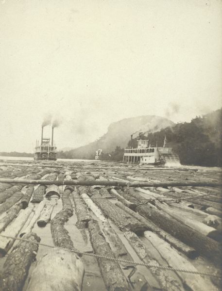 Lumber raft in the water near the shoreline with two paddle wheel steamboats and a bluff in the distance.