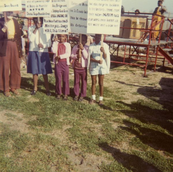Three children stand holding signs along with adults. They're protesting city neglect of the Ninth Ward in New Orleans. In the background are musicians on a stage.