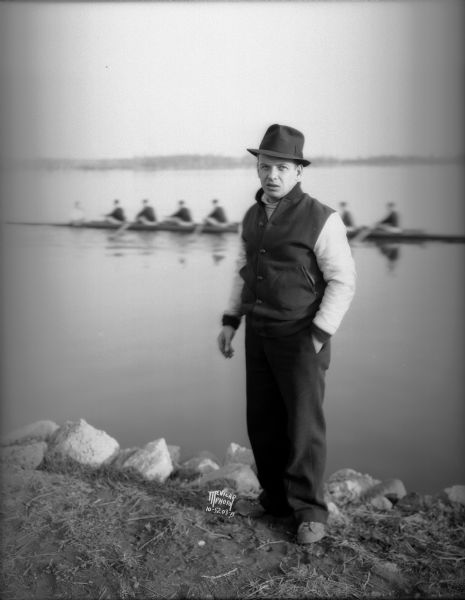 Portrait of Ralph Hunn, coach of the University of Wisconsin-Madison crew standing on the shoreline of Lake Wingra. In the background the crew is rowing on the lake.
