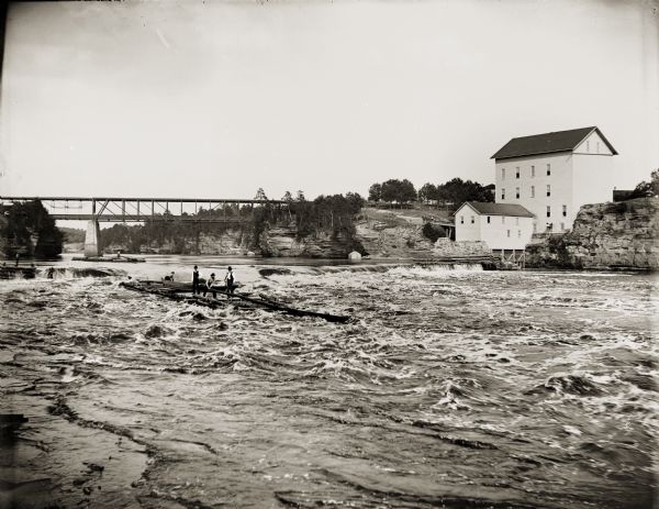 View across water of several men steering a lumber raft over the dam at Munger's Mill. A second raft is in the background, in front of railroad bridge.
