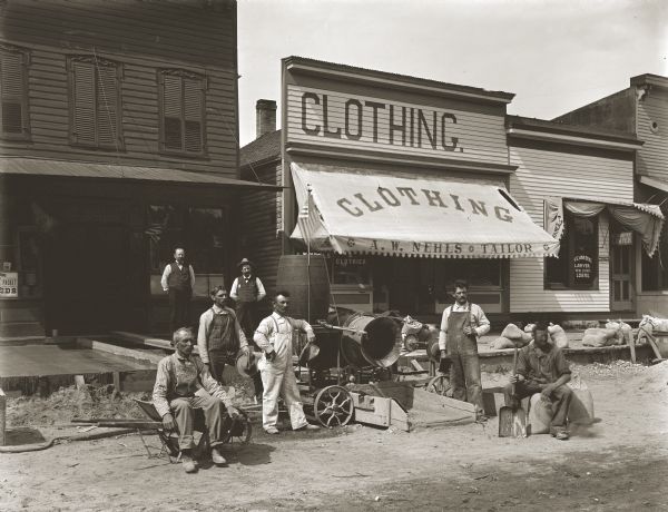 A group of manual laborers posing for a portrait in front of a tailor's shop. The men apparently work pouring concrete to make sidewalks and are posing with the tools of their trade.