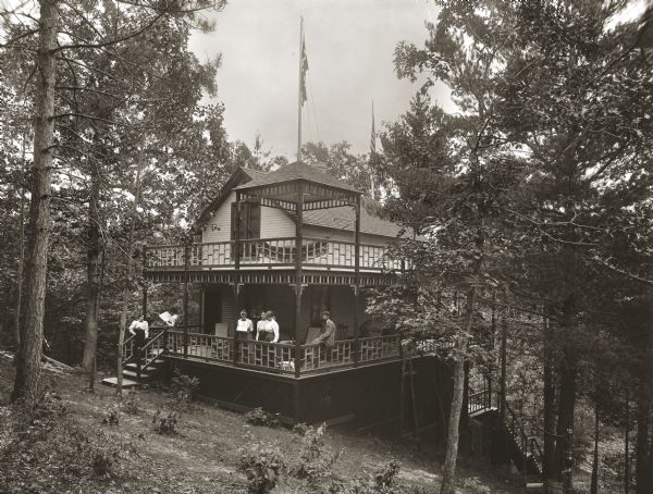 Exterior view looking downhill towards Lark's Hotel surrounded by trees. A group of people and a dog are posing on the porch and front stairs. On the second floor porch, a hammock is strung beneath a small pavilion.