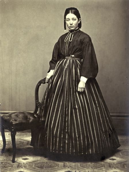 Frankie Irene Douty poses for her wedding portrait. She is wearing a dark dress with a striped skirt and resting her hand on the back of a chair. She has a pocket watch tucked into the waistband of her skirt.