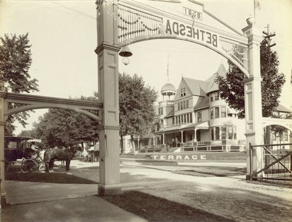 Exterior view of the Terrace Hotel through its front gate, upon which the word "Bethesda" appears backwards. There is a horse-drawn carriage off to the side.