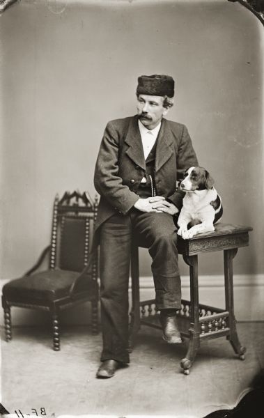 A studio portrait of H.H. Bennett sitting on a small table with a dog. Bennett is wearing a fur hat.