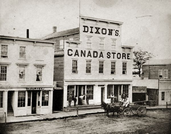 Elevated view of storefront of Dixon's Canada Store. Dixon’s Canada Store was reputed to be the finest dry-goods emporium west of Milwaukee in the mid 1860s. There are three people in a horse-drawn wagon in front of the building, and men and children stand on the steps near the front windows.