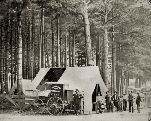 Several men and children pose next to George Houghton's portable photograph tent. There is a wagon parked next to it which bears a sign that reads "Houghton's Photographic Views."