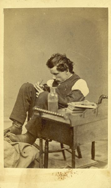 A candid portrait of Henry Hamilton Bennett seated behind a small table. He appears to be touching up a photograph or writing on it. On the table is a glass bottle and a stack of photographs.