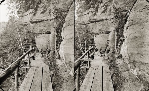 Stereograph of two girls standing on a small wooden bridge with a railing along a rock formation.