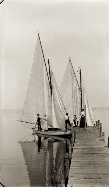 View from shoreline towards a group of people at the end of a pier standing near or on two sailboats. A shoreline is in the far background.