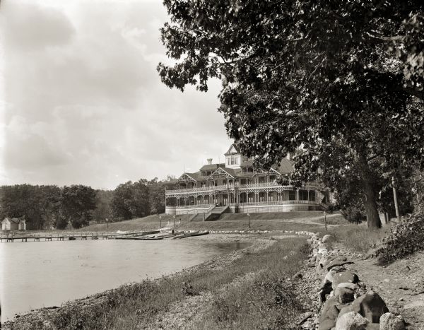 View along shoreline of the hotel with wrap-around porches on the shore of Fox Lake. There are canoes near a pier, and small outbuildings among trees.