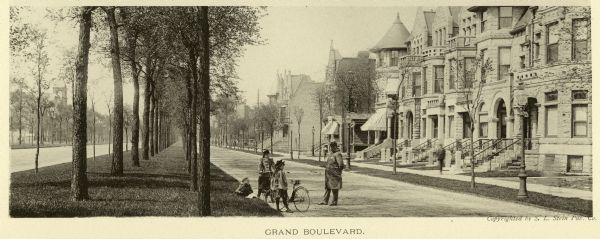View down grassy median planted with trees in the center of Grand Boulevard. Stone residences are on either side. A man, perhaps a police officer, stands in the street talking with three children who have two bicycles.
