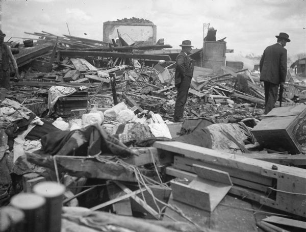 Two well-dressed men survey piles of debris left in the wake of a tornado. Part of the wall of a brick building is in the background.