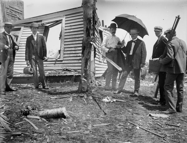 Men, one of whom is holding an umbrella, stand looking at a tree that has been stripped of its bark by a tornado. It also appears to be embedded with a piece of metal. Another man holds a tripod and perhaps a camera case. Behind them are the dilapidated remains of a house.