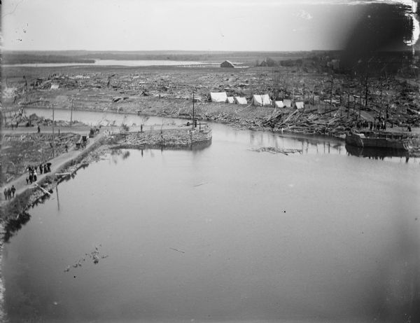 Elevated view over water of the damage created by a tornado. A river, with debris visibly floating in it, runs through the town. Groups of people are walking through the streets, and tents are along the shoreline.