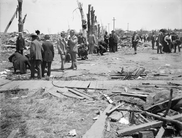 A crowd gathers amid debris left from the aftermath of a tornado. Trees have been stripped of their limbs and bark, and houses have been flattened by the natural disaster.
