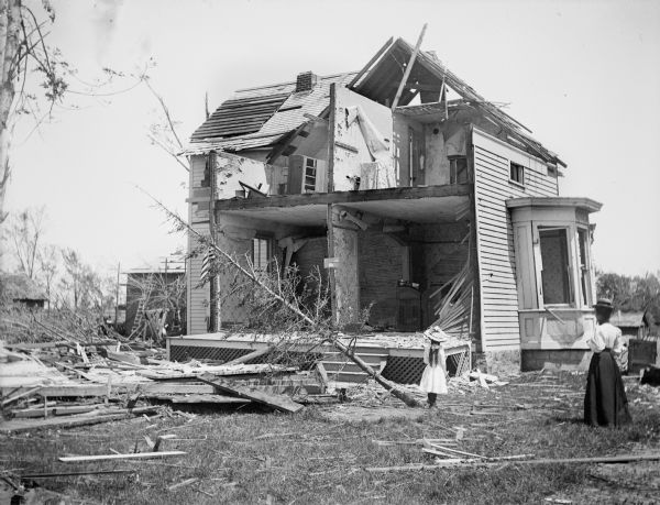 A woman and her young daughter stand in a yard examining a house in the aftermath of a tornado. The house has one entire side missing, a tree leans over on its side, and debris fills the yard.