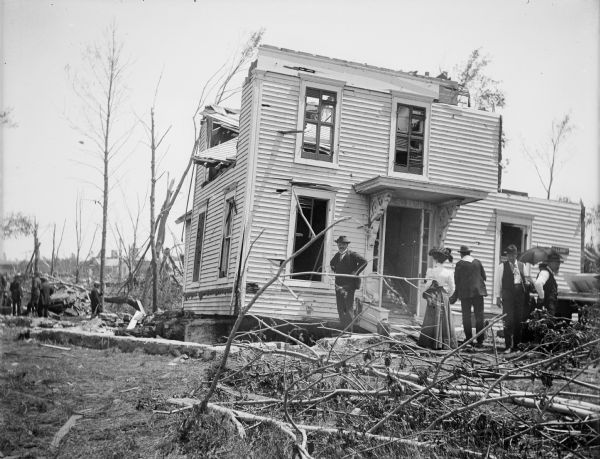 A small group of people stand in the yard of a damaged house that had its roof removed by a tornado, and moved off of its foundation, which is exposed on the left. Debris covers the yard, and trees are stripped of leaves and branches.