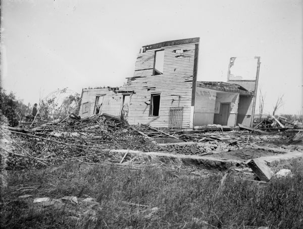The remains of a house damaged by a tornado. The roof and much of the structure have been completely removed and destroyed. Other debris litters the surrounding area, and a man stands on the left.