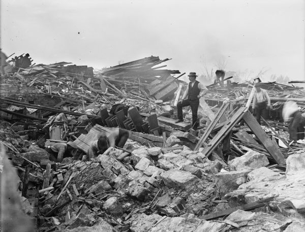 A man wearing a hat stands among piles of rubble left in the wake of a tornado. A number of men are working in the large piles of debris, which includes lumber, bricks, and stone.
