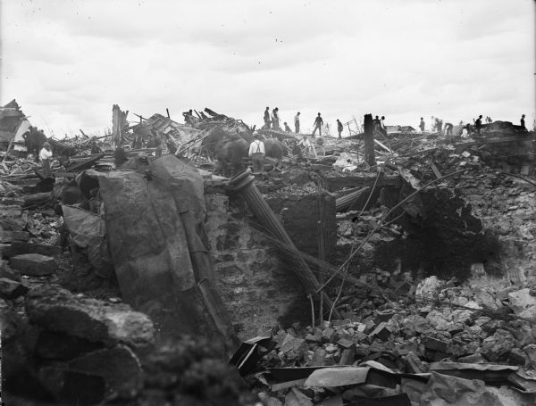 A group of men working in the debris left in the wake of a tornado. A large column is leaning against part of a wall or foundation.
