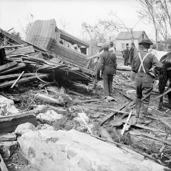 Man standing around the remains of a house that has been demolished during a tornado. A man is standing near a horse on the right. In the background a small structure is damaged but intact.