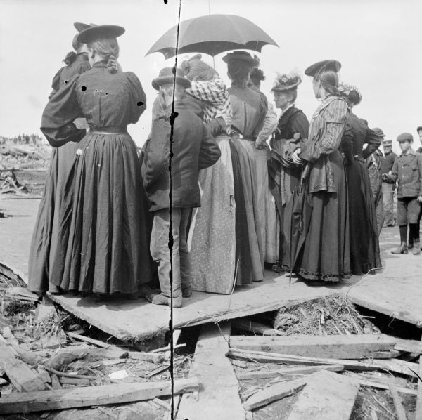 Rear view of a small group of women and children observing the aftermath of a tornado. They are standing on boards, and debris covers the ground.