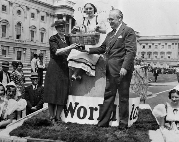 Senator Wiley and Mrs. John Nance Garner, wife of the Vice President, stand on either side of a woman holding a basket and sitting on the world's largest cheese on a parade float.