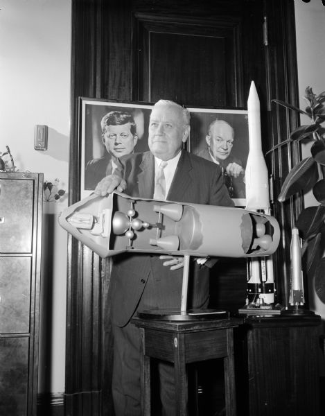 Senator Alexander Wiley posing with a cutaway model of an Apollo rocket. Behind him on a wall are portraits of Dwight D. Eisenhower and John F. Kennedy.