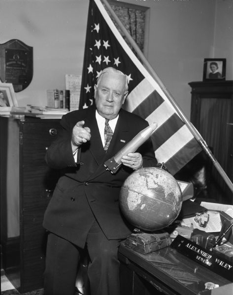 Senator Alexander Wiley posing with a model of a missile. Near him on the desk are an American Flag and a globe.