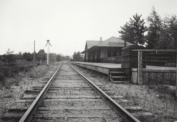 View down railroad tracks which pass the Miscauno Island depot. A railroad signal is on the left, and the platform for the depot is on the right.