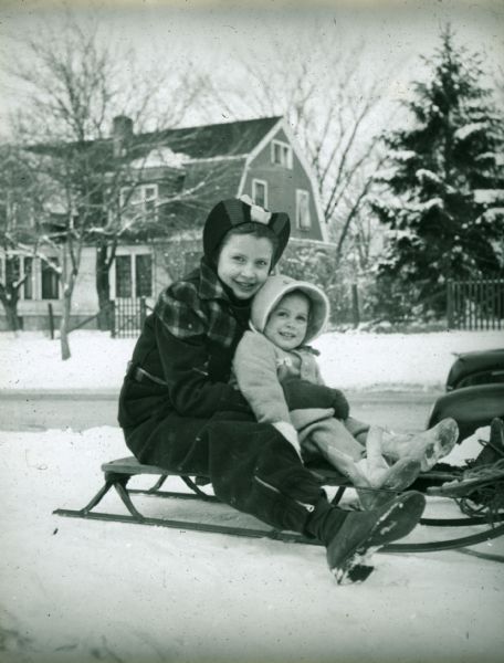 Winter scene with two children on sled in the snow, possibly on Regent Street, Madison, Wisconsin. In the background is a house and yard.