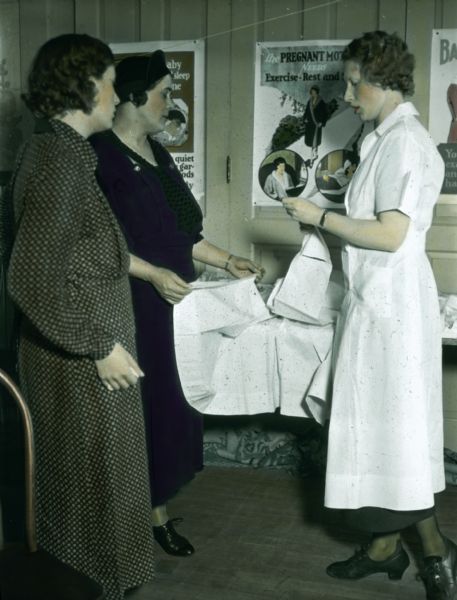 A nurse is showing expectant mothers an exhibit. including maternity clothes. Posters on health are displayed on the wall behind them.