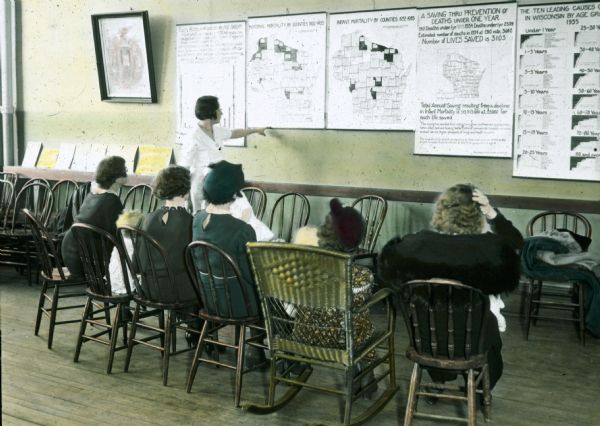 View from behind of group of woman and children sitting in chairs and listening as an instructor points to a chart on the wall. The instructor is teaching women about health.