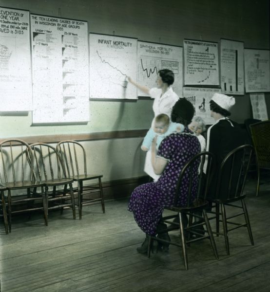 A nurse points to charts while teaching maternal and infant health to a small class of mothers who have children on their laps.