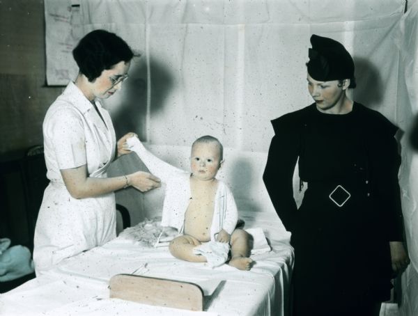 A nurse undresses a baby in preparation for a physical examination as the fashionably-dressed mother looks on.